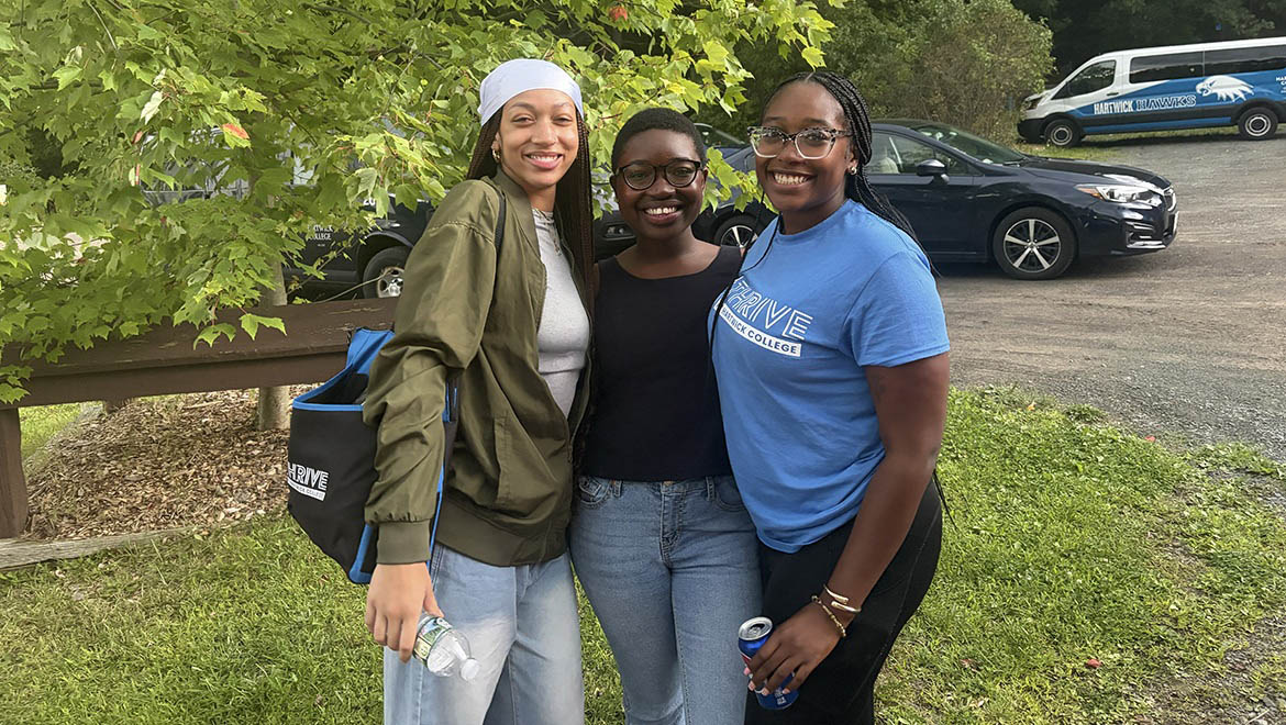 Three smiling young women stand outdoors. One wears a white headscarf and green jacket, another a black top, and the third a blue 'Thrive Hartwick College' t-shirt. A van with 'Hartwick Hawks' is in the background.
