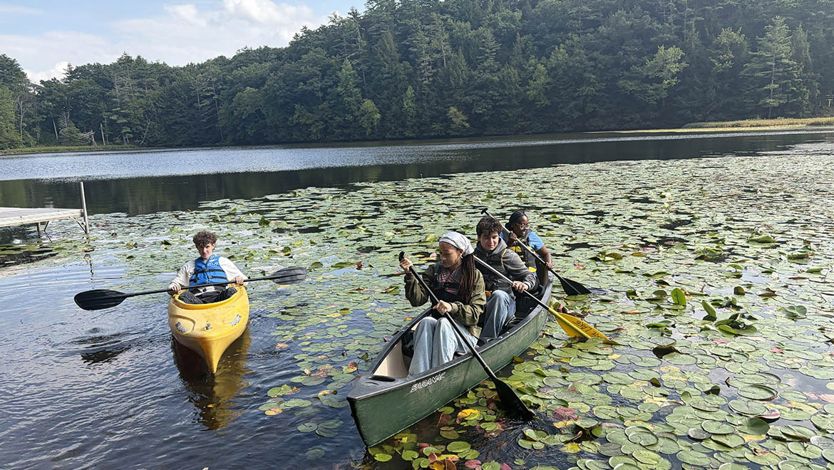 Four Hartwick College students in a kayak and a canoe on a lake covered with lily pads, with a forest in the background.