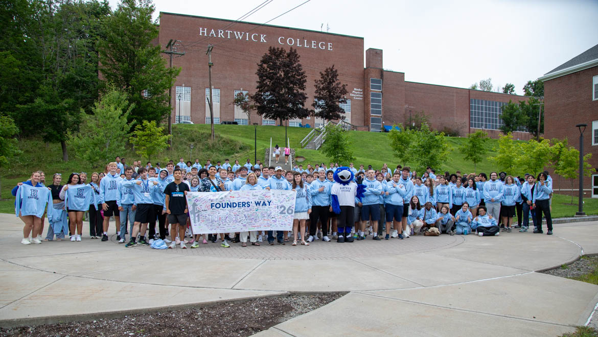 A large group of students and staff, many wearing light blue Hartwick College hoodies, stand on a paved area in front of the Hartwick College building. They hold a banner reading 'Founders' Way First Walk '26'. The college mascot, a blue bird- Swoop, is among them.
