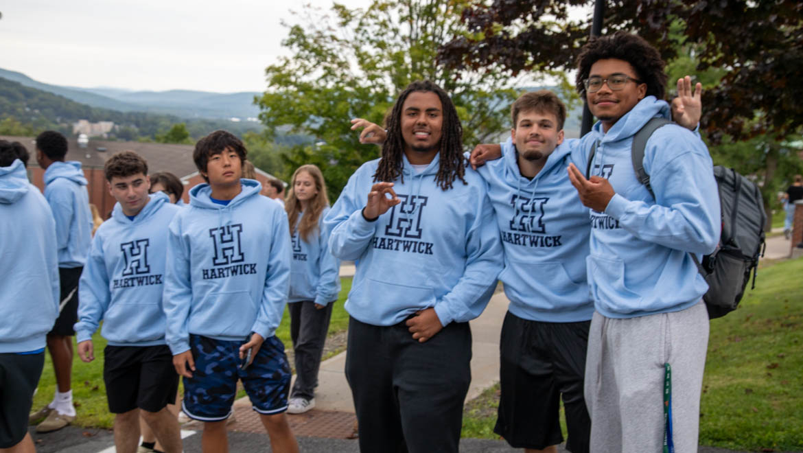 Hartwick new students during First Walk wearing light blue hoodies with 'H HARTWICK' logo, posing outdoors.