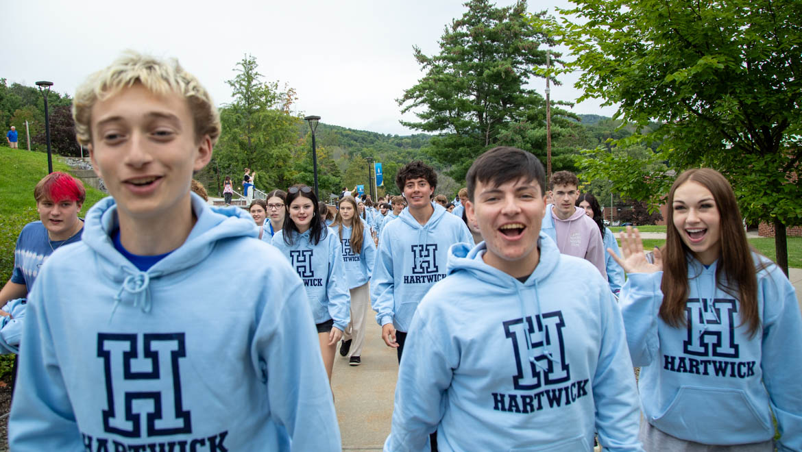 A group of smiling new Hartwick students, mostly wearing light blue Hartwick hoodies, walk outdoors on a campus path during First Walk