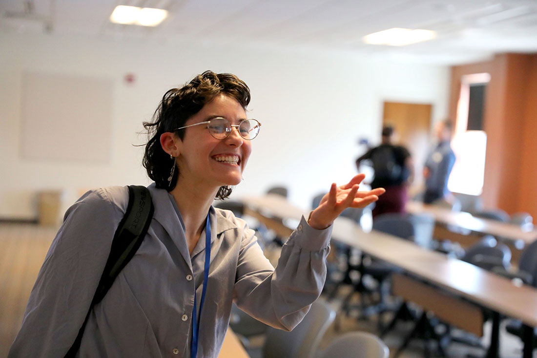 A person with short, curly dark hair and glasses smiles and gestures with an open hand in a classroom.