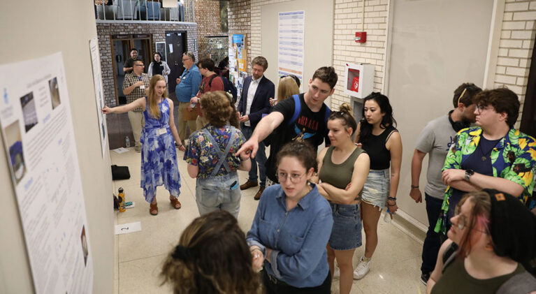 A group of people, mostly young adults, gathered in a hallway, looking at posters and conversing at an event.