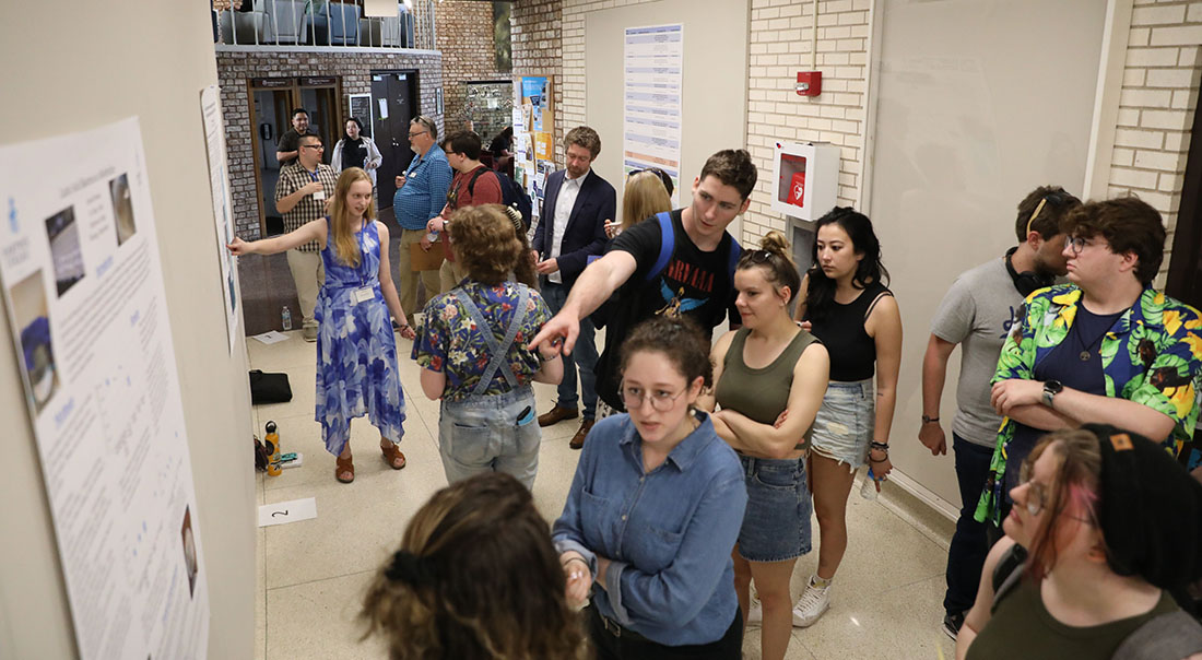 A group of people, mostly young adults, gathered in a hallway, looking at posters and conversing at an event.