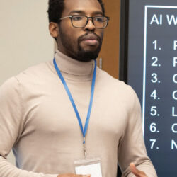 A man in a beige turtleneck and glasses presents next to a screen displaying an 'AI W' list.