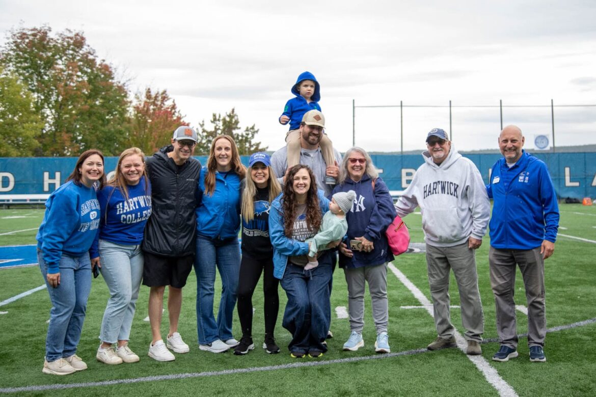 A large group of adults, a child, and a baby pose on a green sports field, many wearing blue Hartwick College apparel.