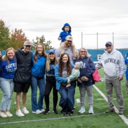 A large group of adults, a child, and a baby pose on a green sports field, many wearing blue Hartwick College apparel.