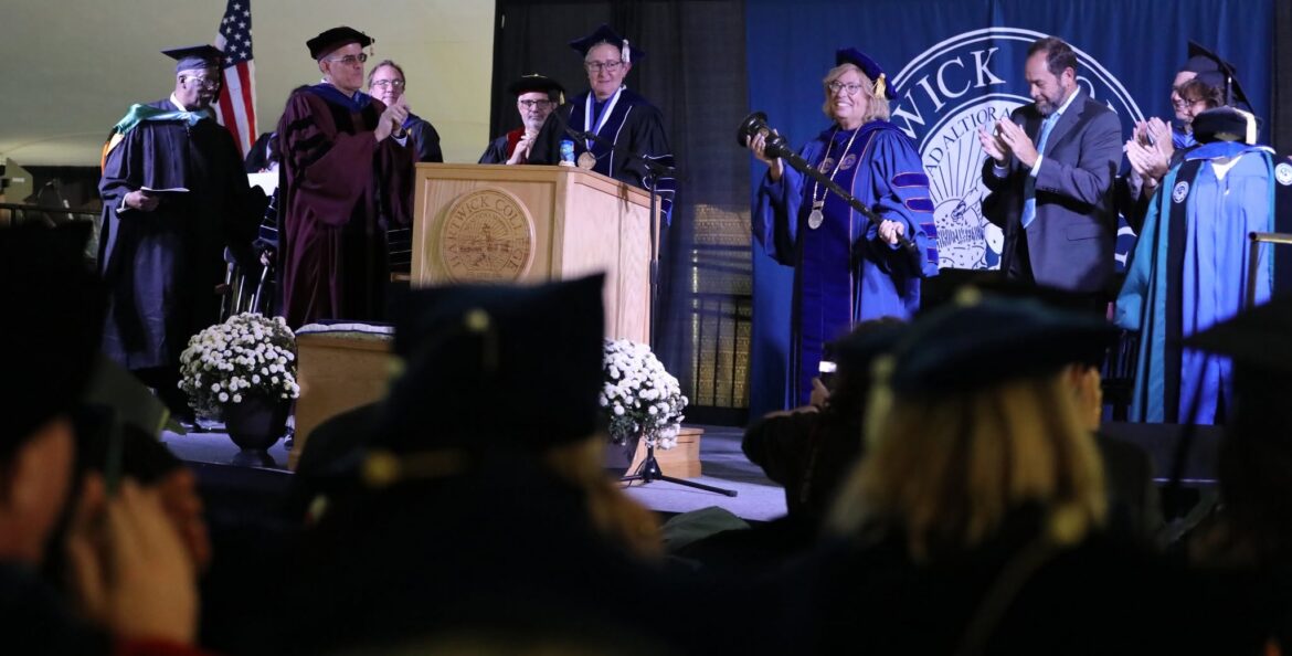 An academic ceremony with several people in academic gowns and caps on a stage. A woman in blue regalia holds a ceremonial mace. A podium with 'Hartwick College' is in the center, and an American flag is on the left. Audience members are blurred in the foreground.