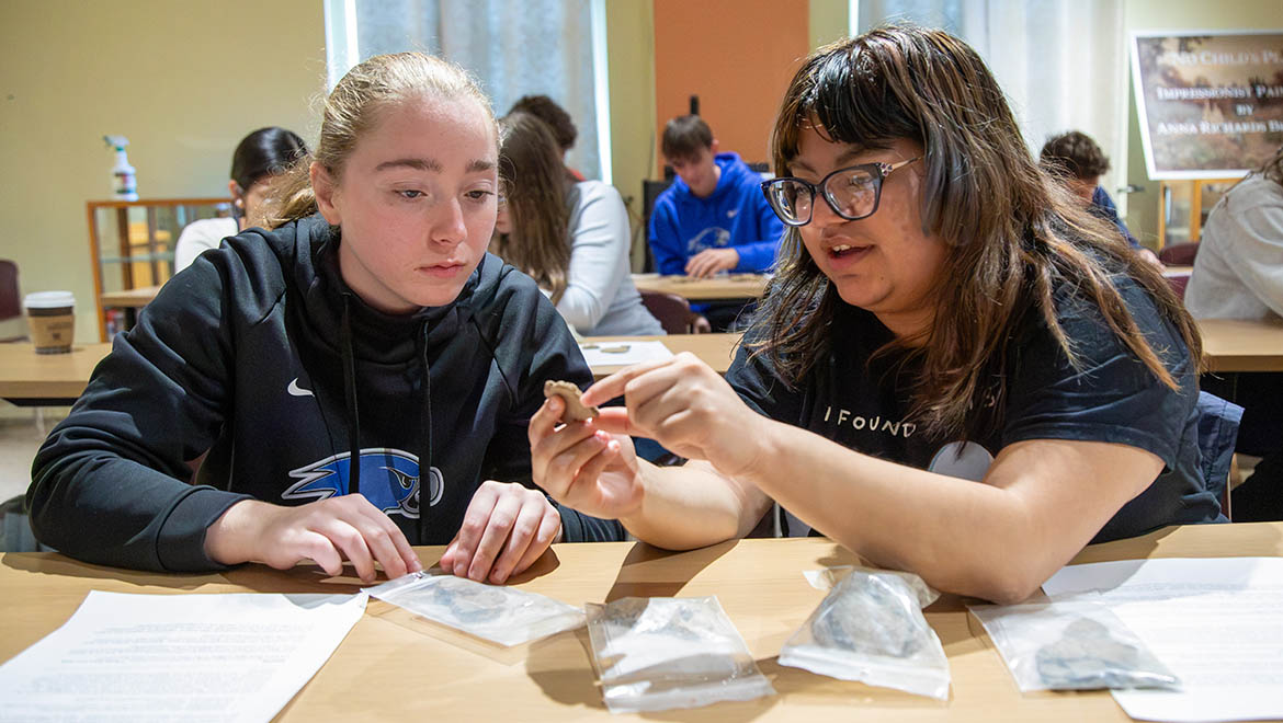 Two young women at a table examine a small, irregular object. One holds it while the other looks on intently. Plastic bags with similar objects and papers are on the table.