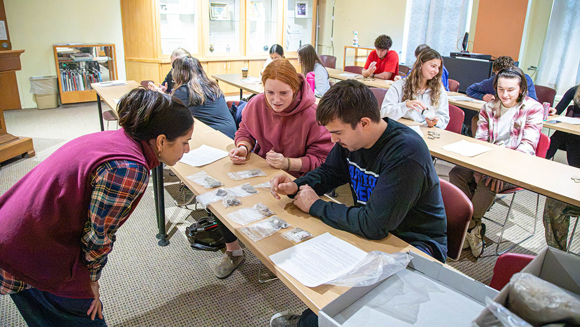Students in a classroom examine small objects in plastic bags on wooden tables, with a woman in a maroon vest leaning in to observe.