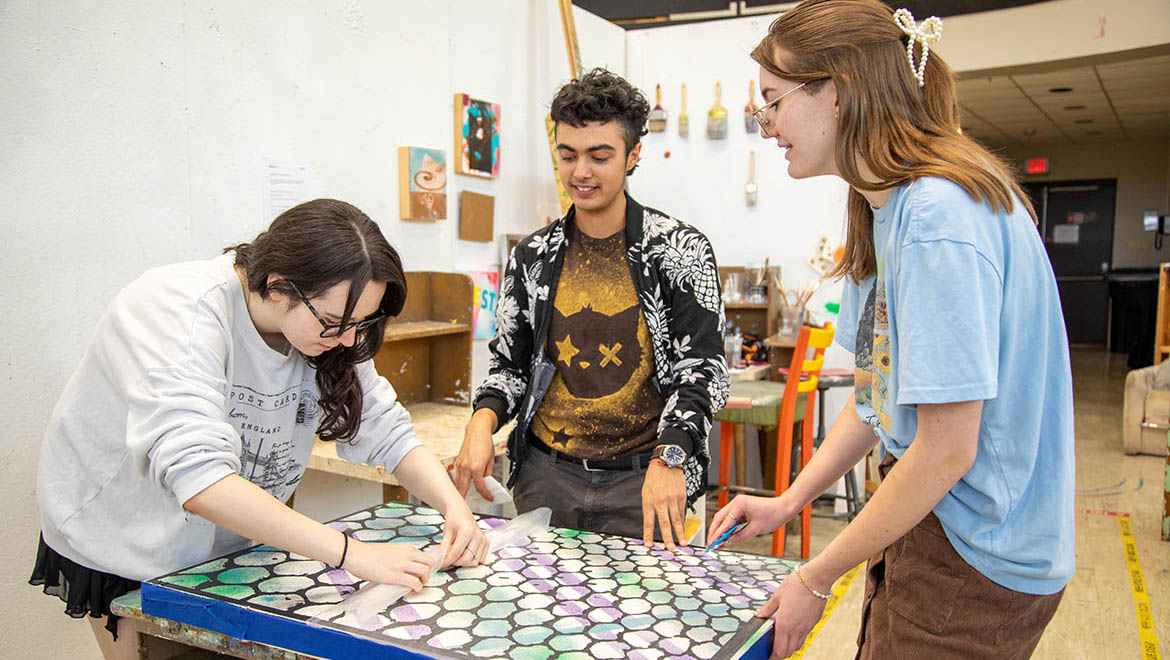 Three young artists, two females and one male, work together on a large art piece with a colorful hexagonal pattern on a table in an art studio.
