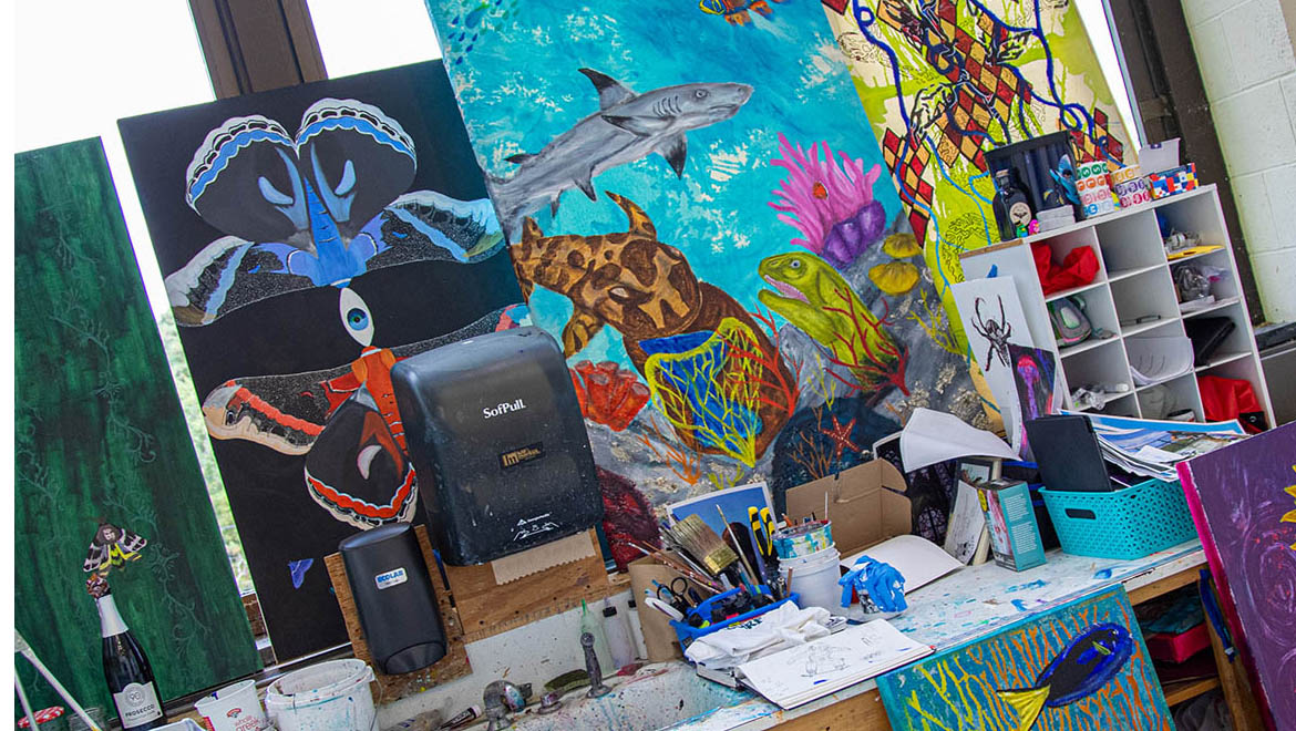 A cluttered art studio with multiple colorful paintings, including an underwater scene, moth wings, and abstract designs, alongside various art supplies on a paint-splattered desk.