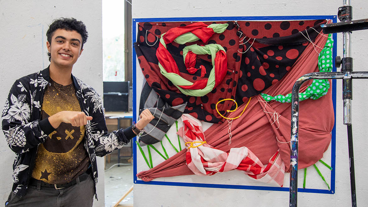 A smiling young man in a patterned jacket points to a vibrant fabric art piece with red, black, green, and white textiles.