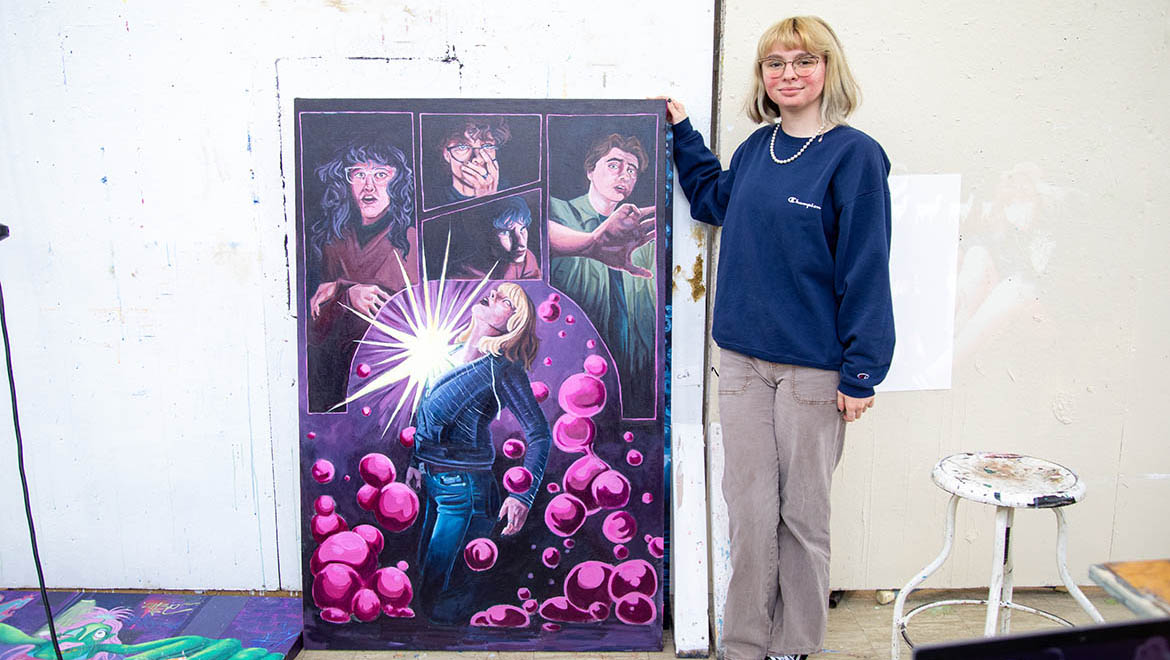 A young woman with blonde hair and glasses stands beside her large, colorful painting depicting a dramatic comic book scene with a central figure glowing and surrounded by pink orbs, while four male figures look on in shock from panels above.