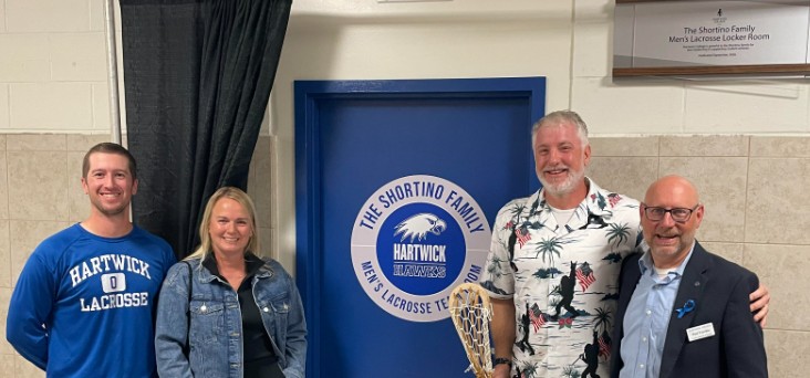Four people smiling in front of a blue door for 'The Shortino Family Men's Lacrosse Team Room' and a plaque for the Men's Lacrosse Locker Room.