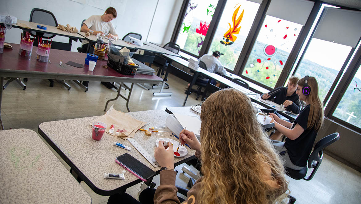 Students in an art classroom, some painting and drawing, with large windows decorated with colorful art and overlooking green hills.