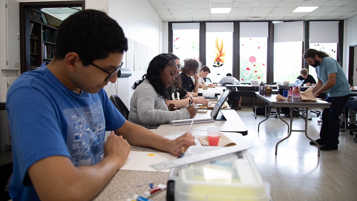 A group of people, including a young man painting and a young woman writing, are engaged in an art workshop in a bright room with decorated windows.