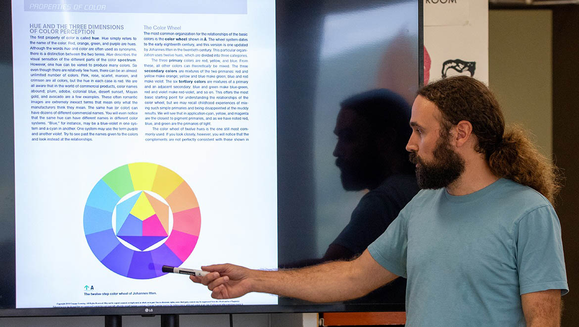 A man with a beard and long hair points at a large screen displaying a color wheel and text about color theory.