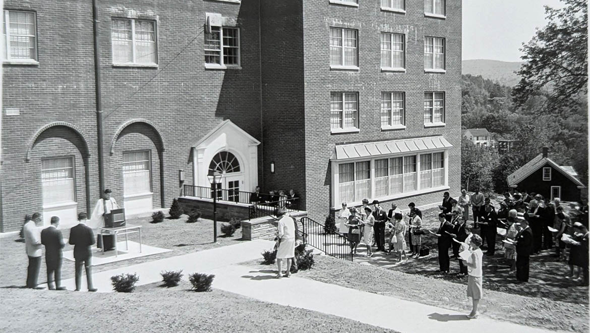 Black and white photo of an outdoor ceremony with a clergyman and attendees in front of a large brick building and wooded hills.