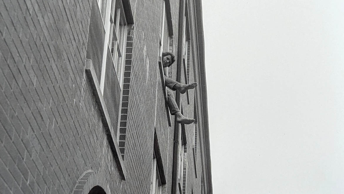 Black and white photo of a person with curly hair and bare feet leaning out of a brick building window.