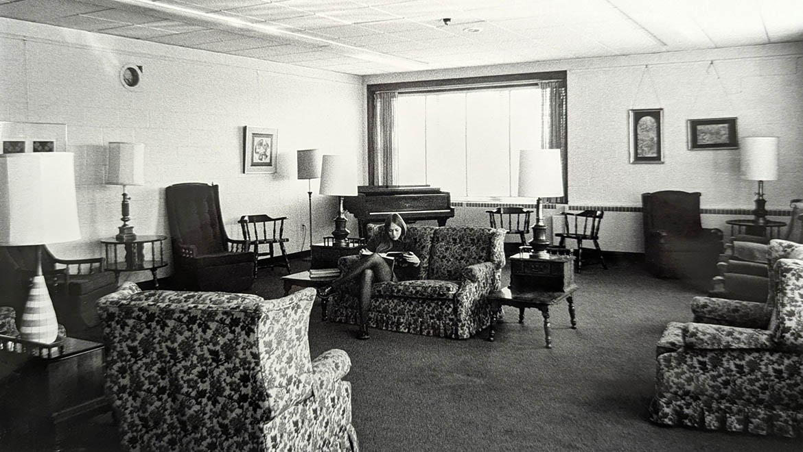 Black and white photo of a woman reading on a floral sofa in a vintage lounge with a piano and multiple armchairs.