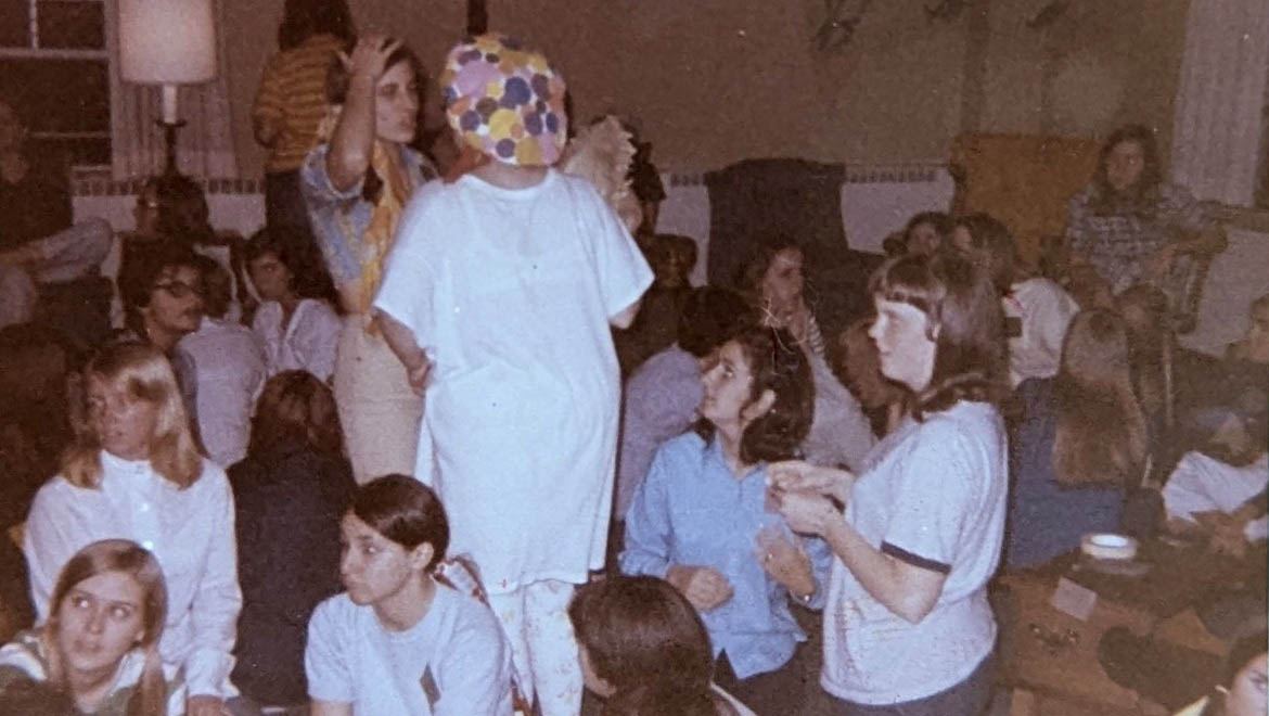 Vintage photo of a crowded indoor social gathering of young people, one person in a white shirt and colorful polka-dot hat, others seated and standing.
