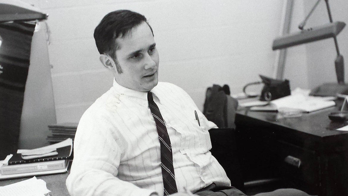 Black and white photo of Hartwick Professor Wendell Frye in a shirt and tie, sitting at an office desk with papers and a lamp.