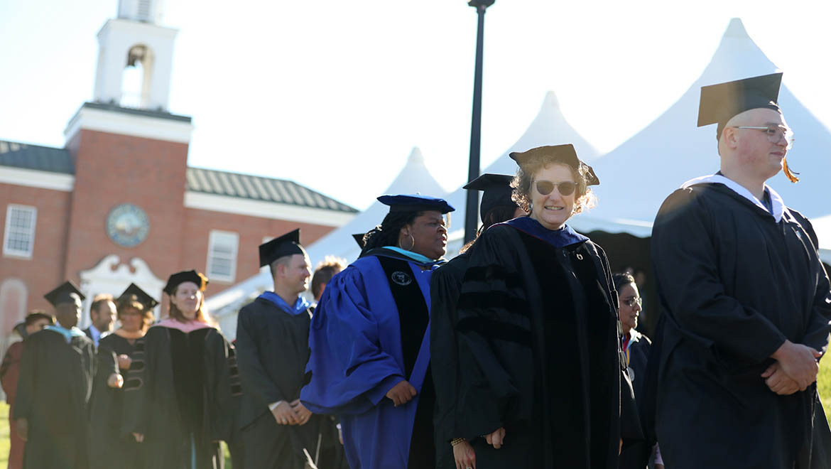 A line of people in academic gowns and caps walking outdoors, with a brick building and white tents in the background.