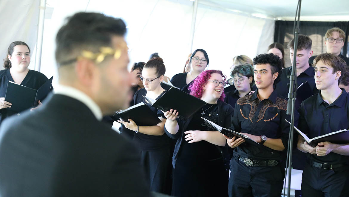 A choir of young adults in black attire sings, holding sheet music. A conductor in a black suit and gold laurel headpiece is in the foreground, seen from behind.