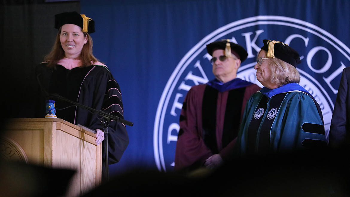 Three people in academic gowns and caps on a stage. A woman at a wooden podium smiles, speaking into a microphone. Behind her, a man in a dark red gown and sunglasses stands next to a woman in a teal gown.