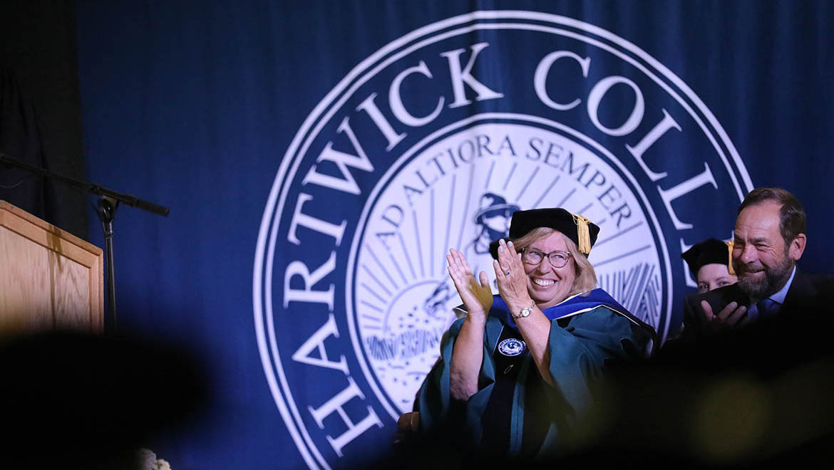 A smiling woman in academic regalia claps in front of a blue Hartwick College banner, next to a smiling man.