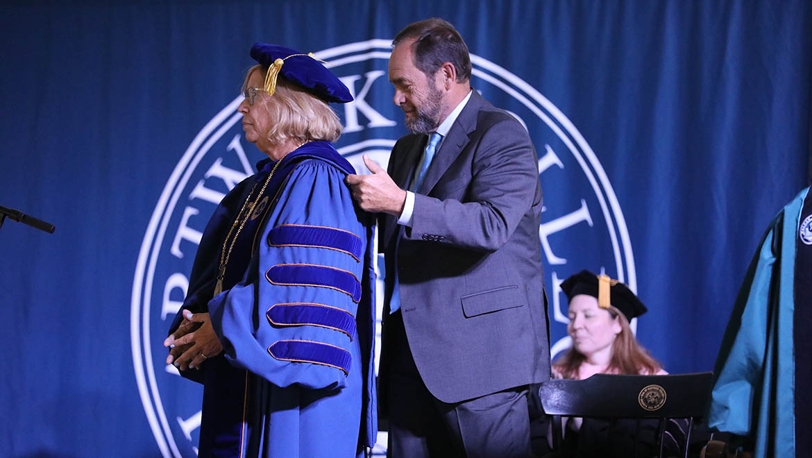 A man in a suit adjusts the academic hood on a woman in blue academic regalia, including a gown, hood, and tam, during a ceremony. A university seal is in the background.