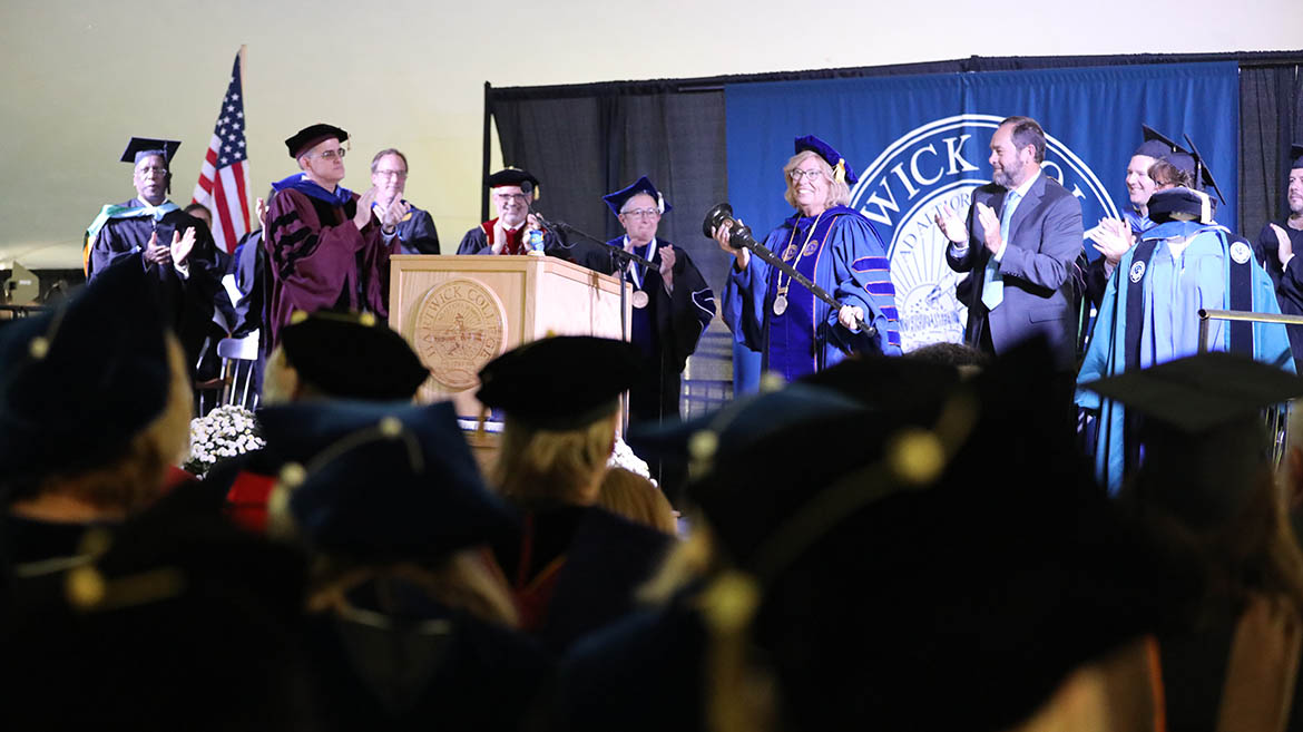 Academic ceremony with faculty on stage clapping, a woman holding a mace, and a Hartwick College banner in the background.