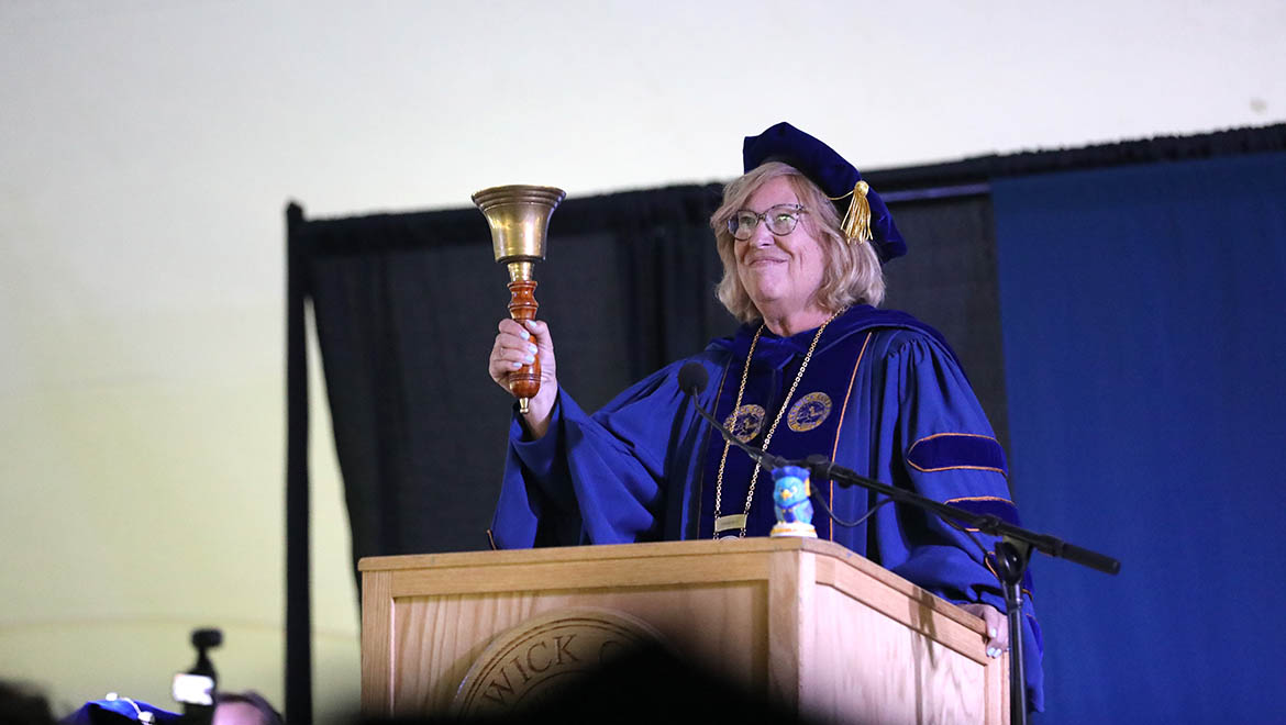 A smiling woman in blue academic regalia and glasses holds a brass bell while standing behind a wooden podium with microphones.