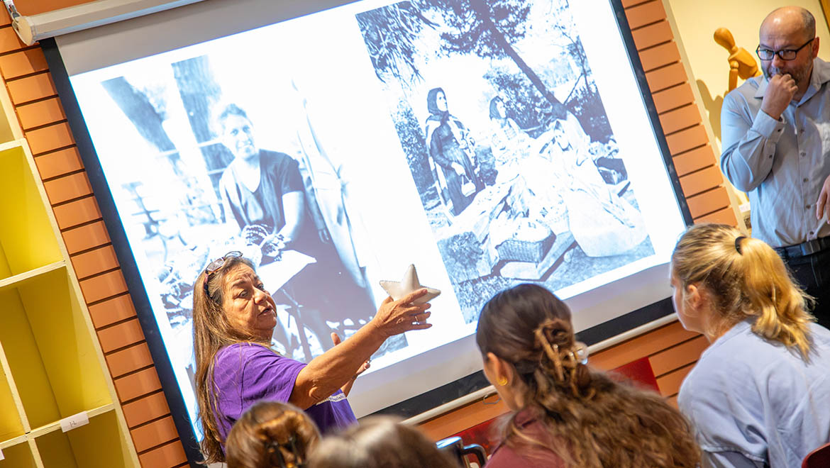 A woman in a purple shirt presents, holding a star, in front of a projector screen showing two black and white photos, with an audience and a man observing.