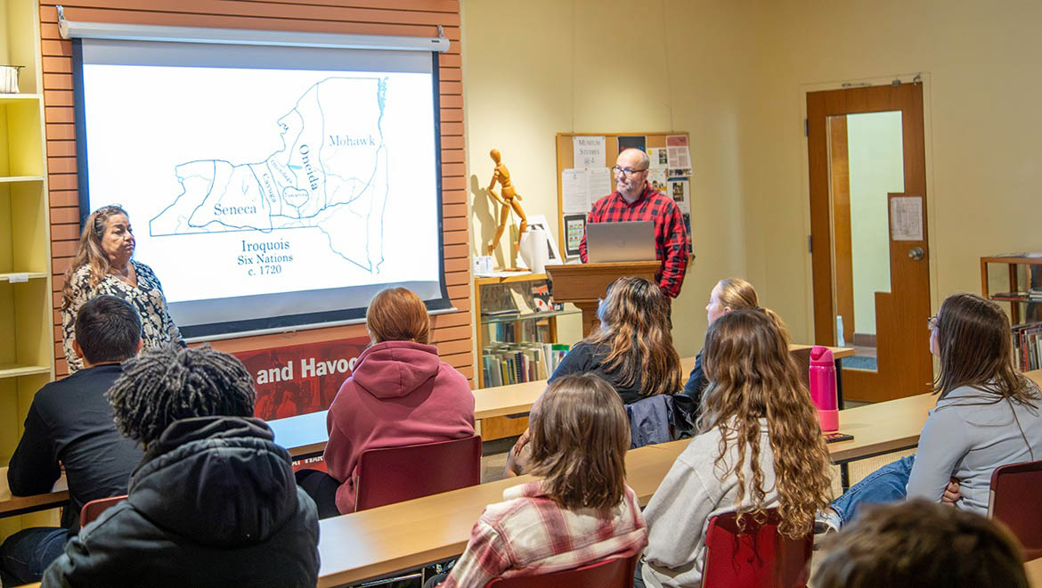 A classroom scene with two presenters and several students. A map of the Iroquois Six Nations is projected on a screen, showing tribal territories in 1720.