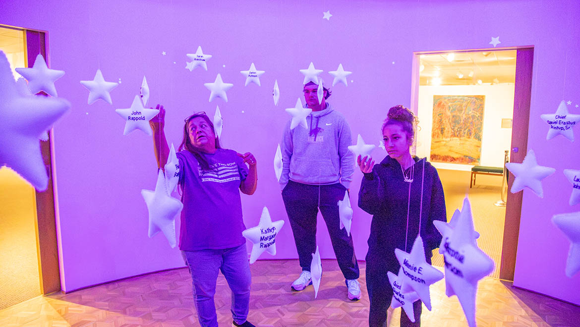 Three people, two women and one man, stand in a purple-lit room surrounded by hanging white star decorations, some with names written on them.
