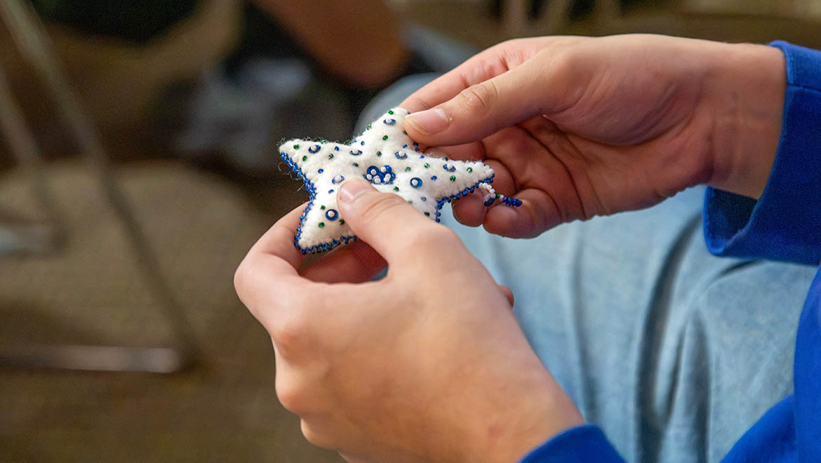 Two hands hold a white, star-shaped craft item decorated with blue and green beads.
