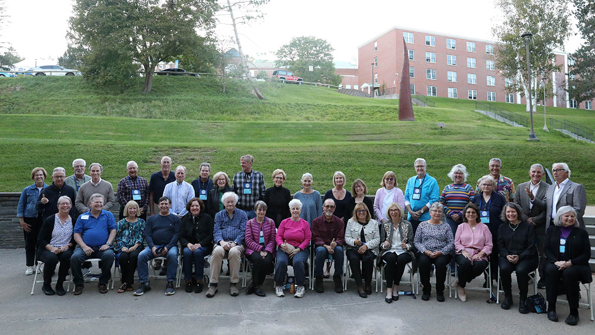 A large group of adults, mostly older, posed in two rows for a photo outdoors. Many wear lanyards. A grassy hill and a brick building are in the background.