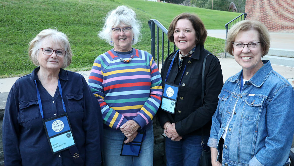 Four smiling women, likely alumni, stand together outdoors, wearing name tags for a 'True Blue Weekend' event.