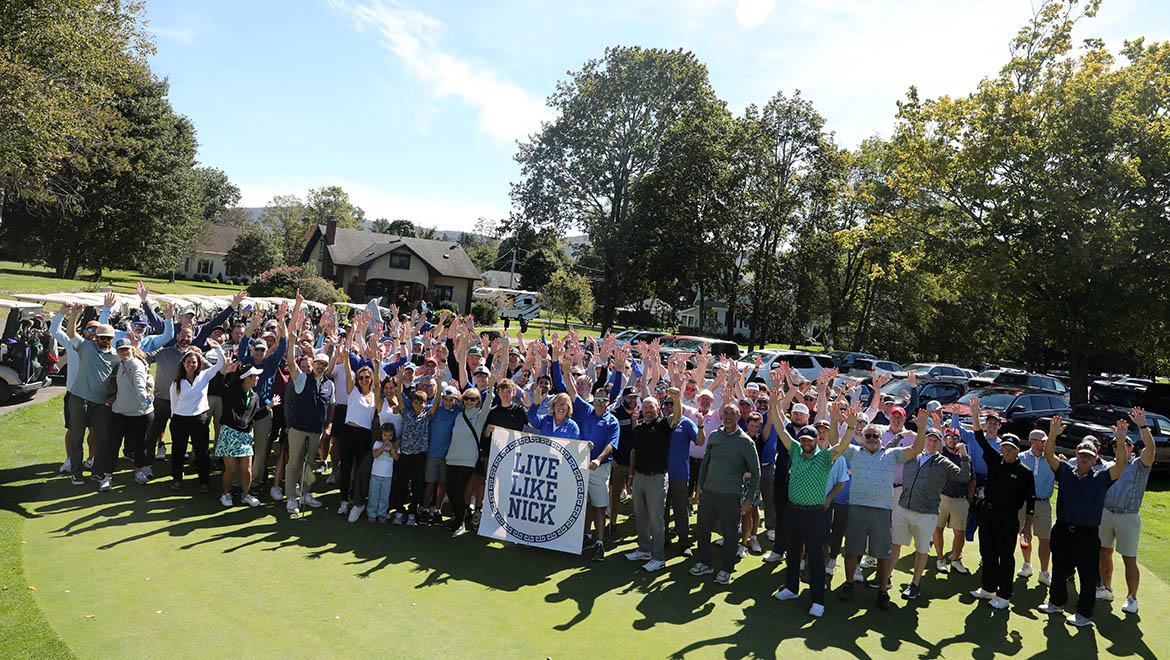 A large group of people on a golf course, hands raised, holding a banner that reads 'LIVE LIKE NICK'.