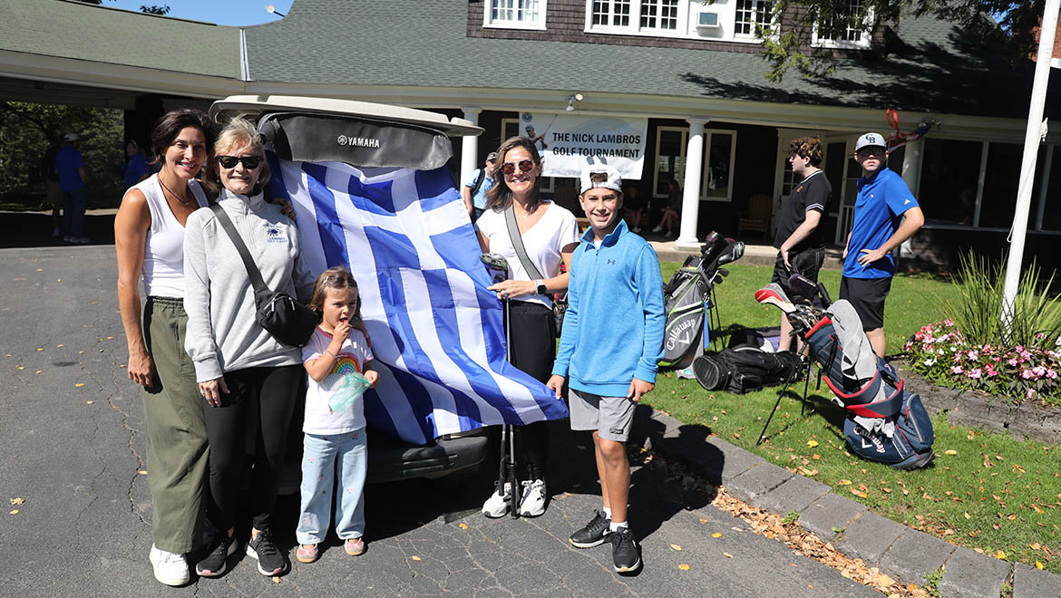 A group of five people, including three women, a boy, and a girl, stand by a golf cart draped with a Greek flag at The Nick Lambros Golf Tournament. Two young men and golf bags are in the background near a clubhouse.