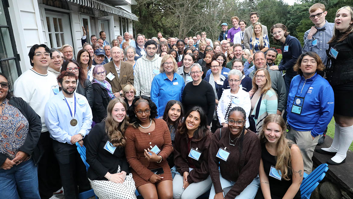 A large group of diverse people of all ages smiling for a photo outdoors in front of a white building with green trees in the background. Many wear name tags, and one person has a medal.