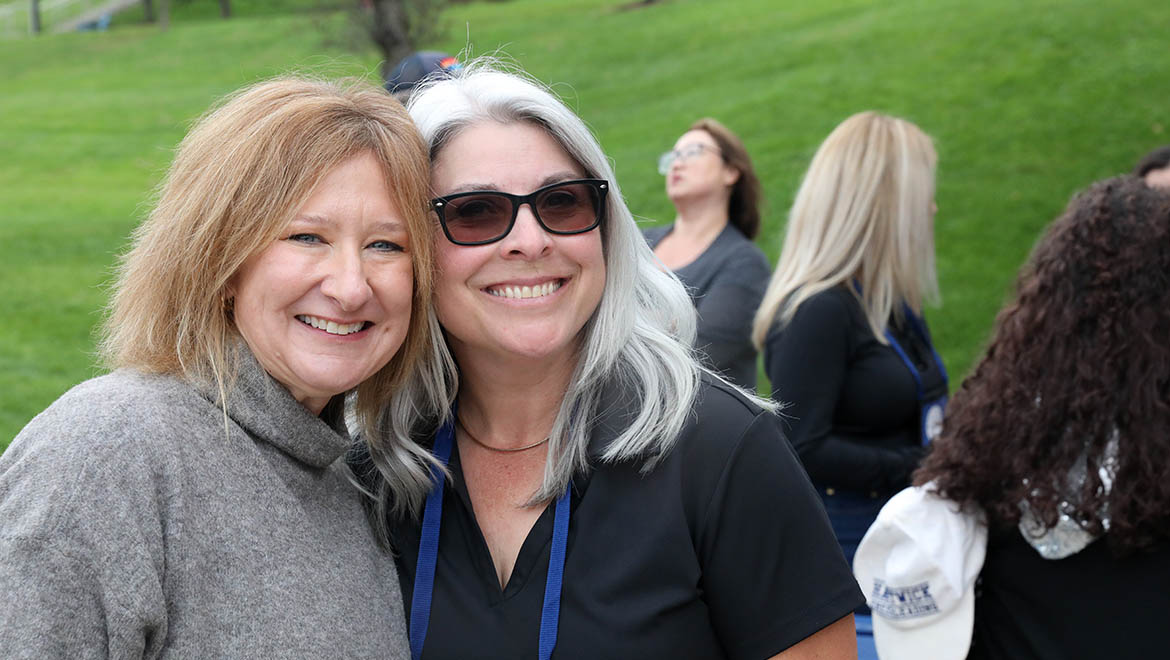 Two smiling women, one with blonde hair and one with grey hair and sunglasses, pose for a photo outdoors with other people in the background.