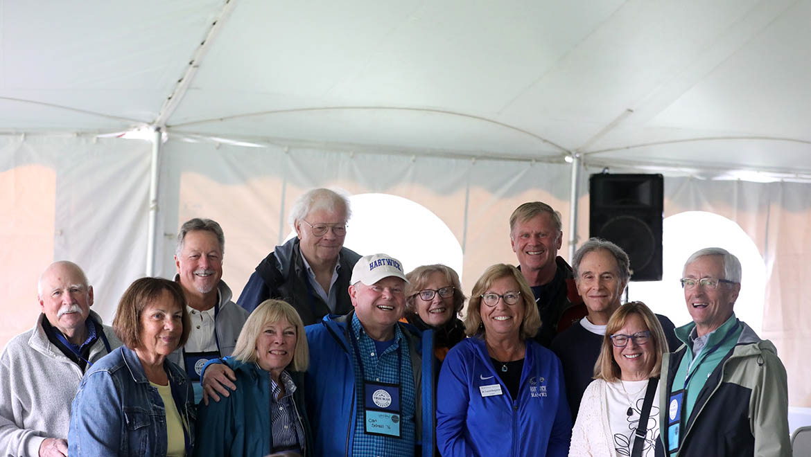 A group of thirteen smiling adults, some wearing Hartwick branded clothing, pose for a photo inside a white tent.