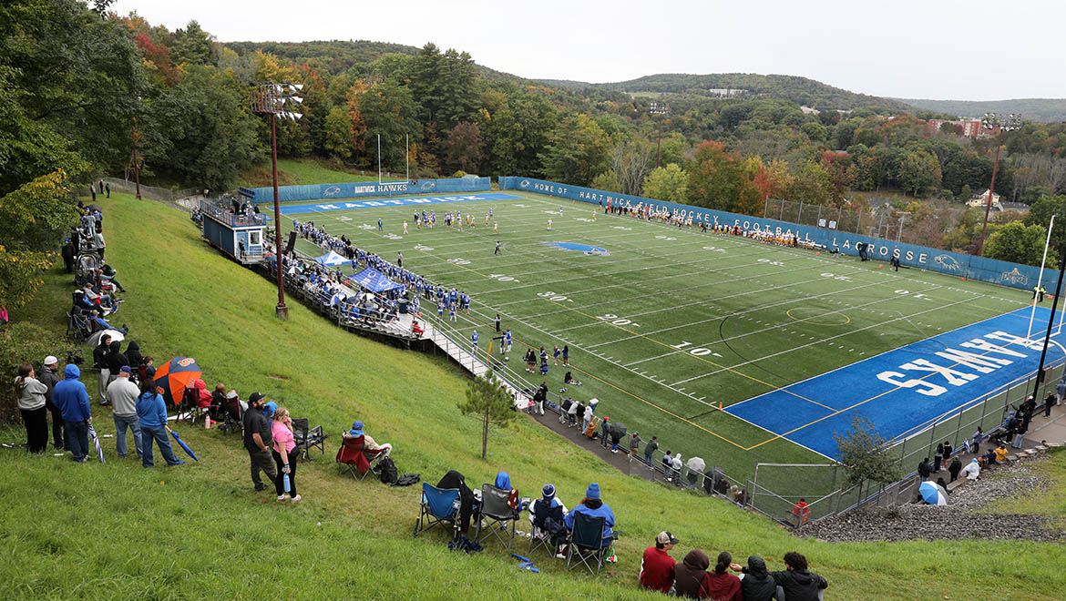 An outdoor sports field with green turf and blue end zones, surrounded by forested hills. Spectators are seated on a grassy slope and in bleachers, watching players on the field under an overcast sky.