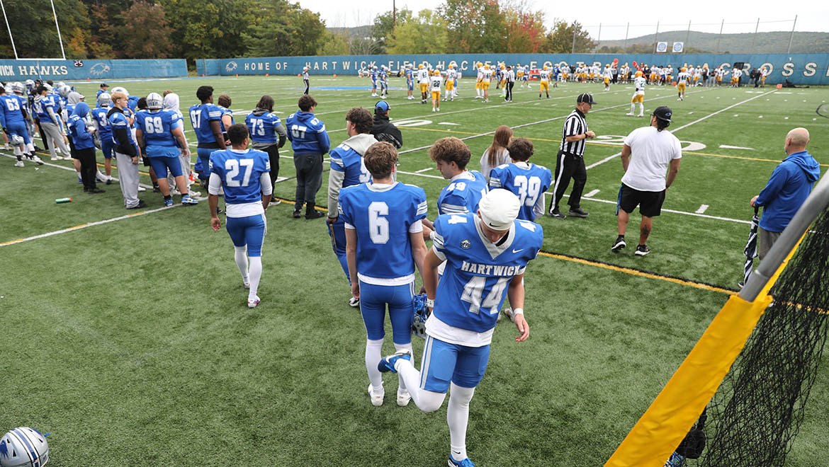 A football field with players in blue and white uniforms, some stretching, and others in yellow uniforms in the distance. A blue fence with 'Hartwick Hawks' text is in the background.