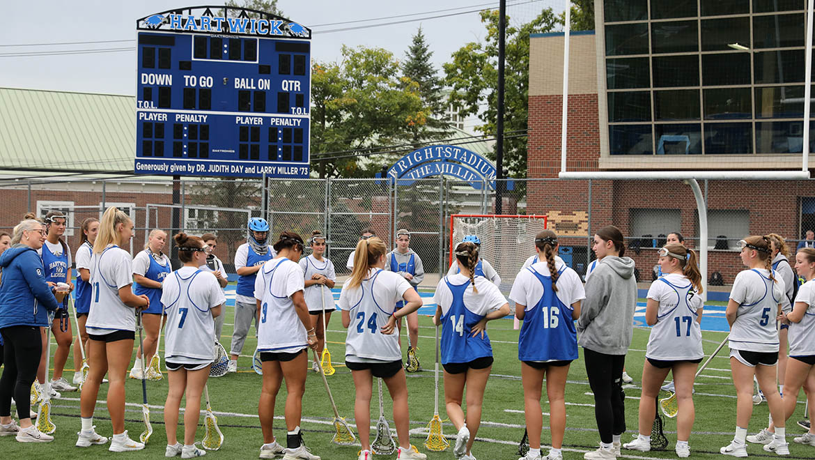 A women's lacrosse team in white and blue jerseys stands on a green field with lacrosse sticks, in front of a blue scoreboard and a stadium sign.