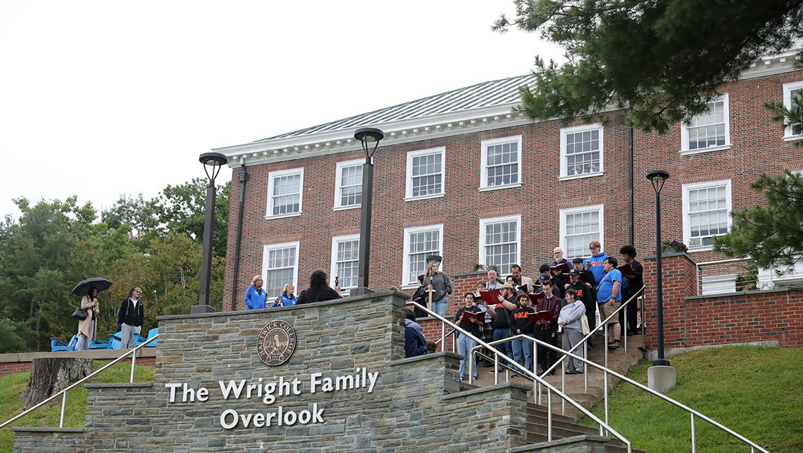 A choir performs on stone steps in front of a red brick building, 'The Wright Family Overlook,' with trees and an overcast sky.