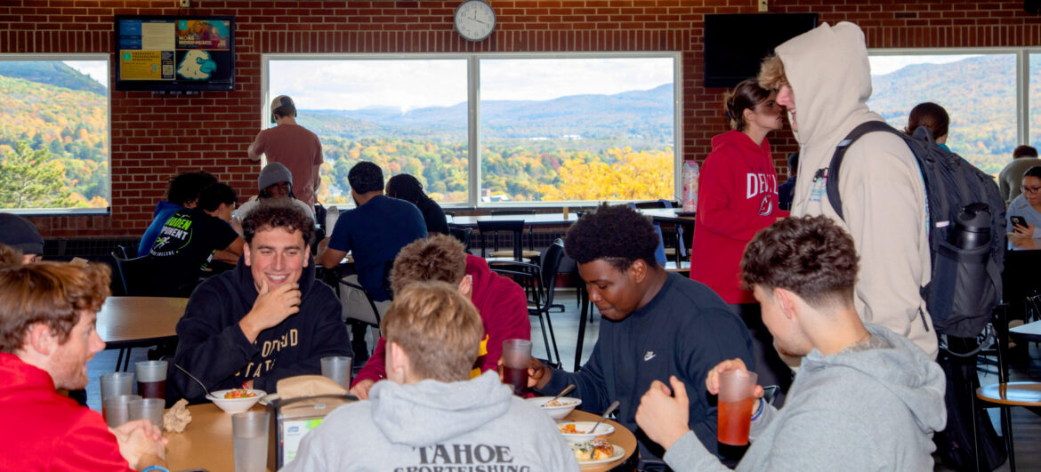 Students eat and socialize in a college dining hall with large windows overlooking a mountain landscape with colorful autumn trees.